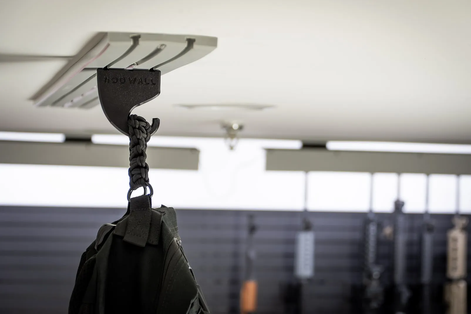 ModWall Ceiling Panel Strip (48″x4″) A close-up view of a black tactical bag hanging from a modular hook on a light grey ModWall Ceiling Panel Strip mounted to a white ceiling. The background shows an organized gun room with additional wall-mounted storage panels and firearms.