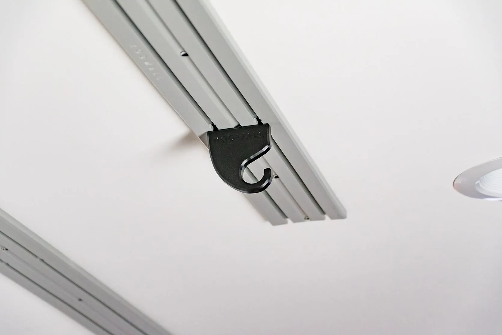 ModWall Ceiling Panel Strip (48″x4″) A close-up view of a black modular hook accessory attached to a light grey ModWall Ceiling Panel Strip mounted on a white ceiling, demonstrating how various accessories can be easily integrated for efficient overhead gear organization.