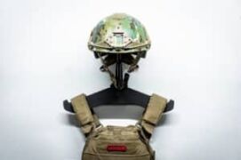 A camo tactical helmet and tactical vest hung on a heavy gear hanger mounted directly to a white wall.
