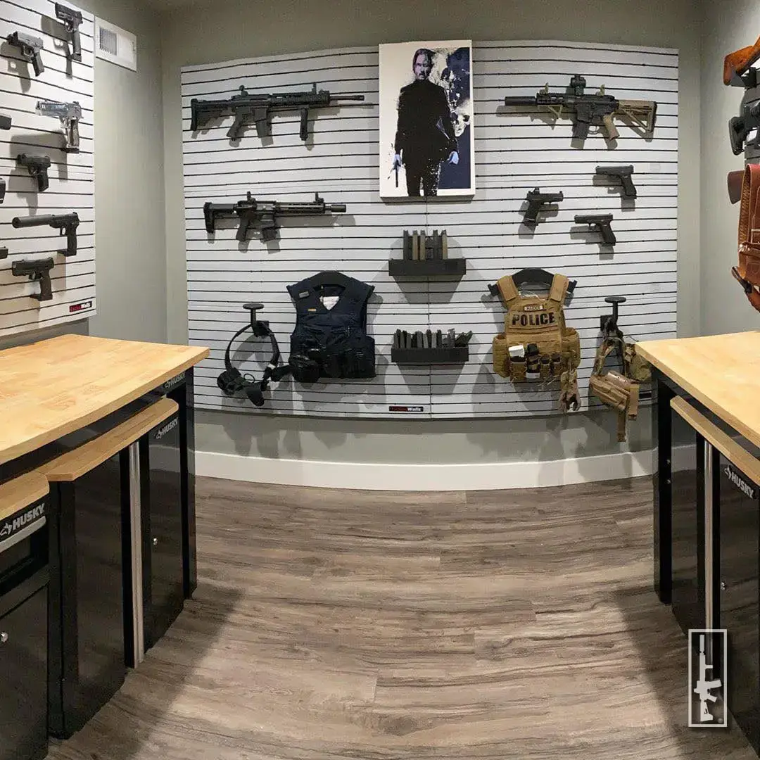 ModWall Panels (48″Wx22″H) A clean vault room utilizing grey ModWall panels to display an array of tactical equipment including horizontally mounted rifles, handguns, and magazine racks. The wall features a central poster, while tactical vests and duty belts are staged on specialized mounts between two professional workbenches.