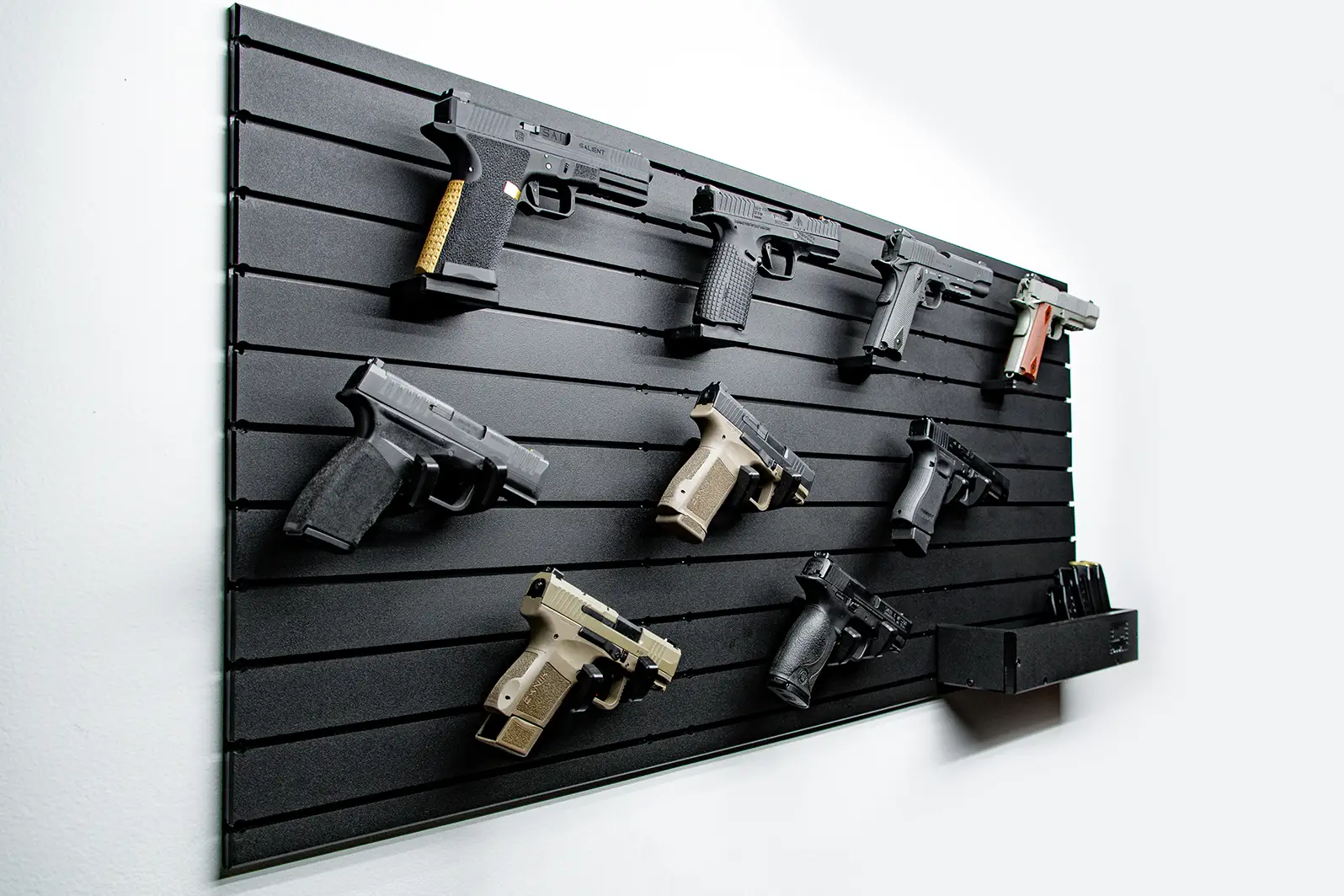 Single Panel Packages A single panel pistol package mounted on a plain white wall, showcasing nine various handguns organized with individual mounts. The modular setup also includes a small shelf at the bottom right for storing magazines.