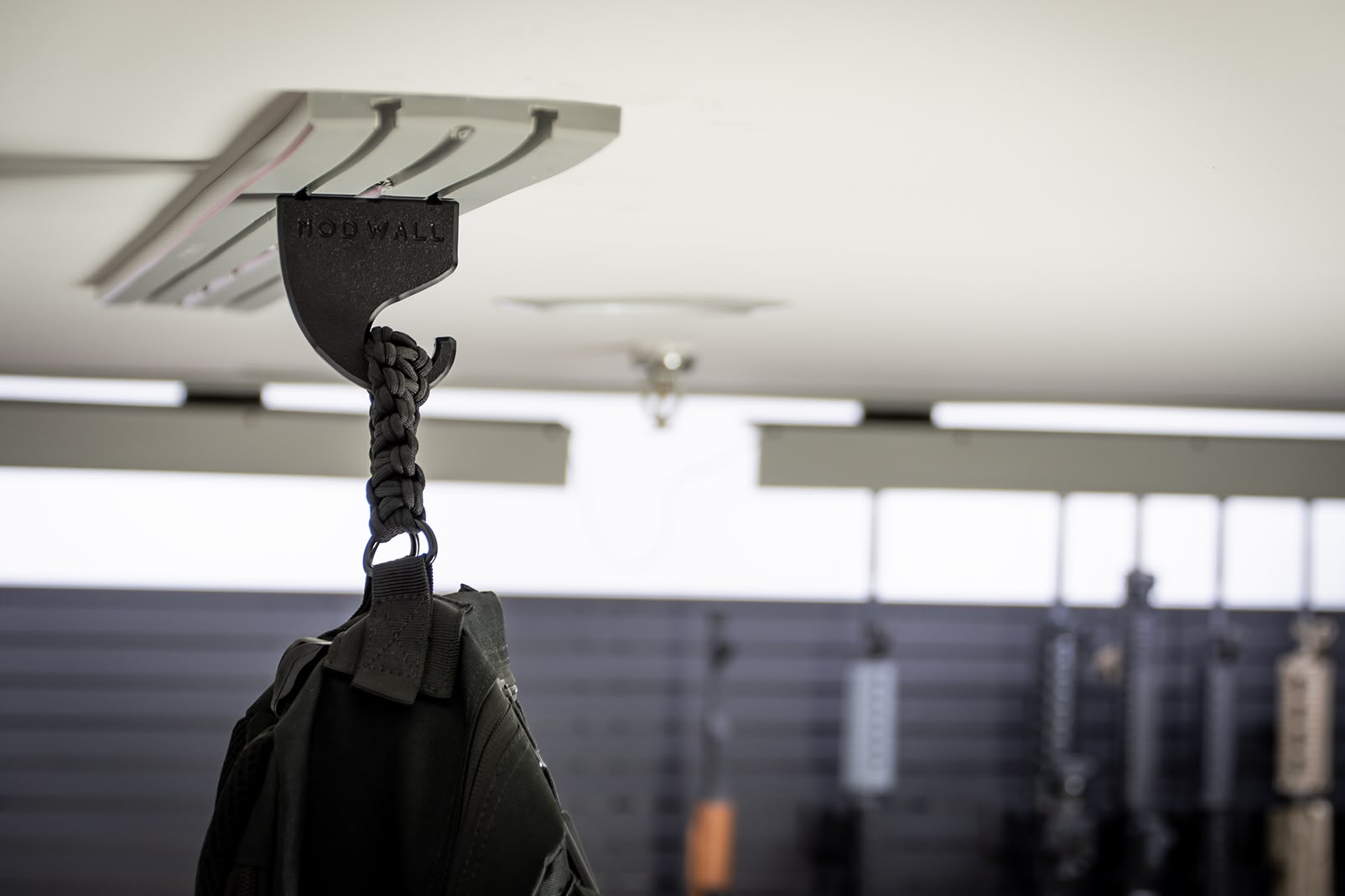 ModWall Ceiling Panel Strip (48″x4″) A close-up view of a black tactical bag hanging from a modular hook on a light grey ModWall Ceiling Panel Strip mounted to a white ceiling. The background shows an organized gun room with additional wall-mounted storage panels and firearms.