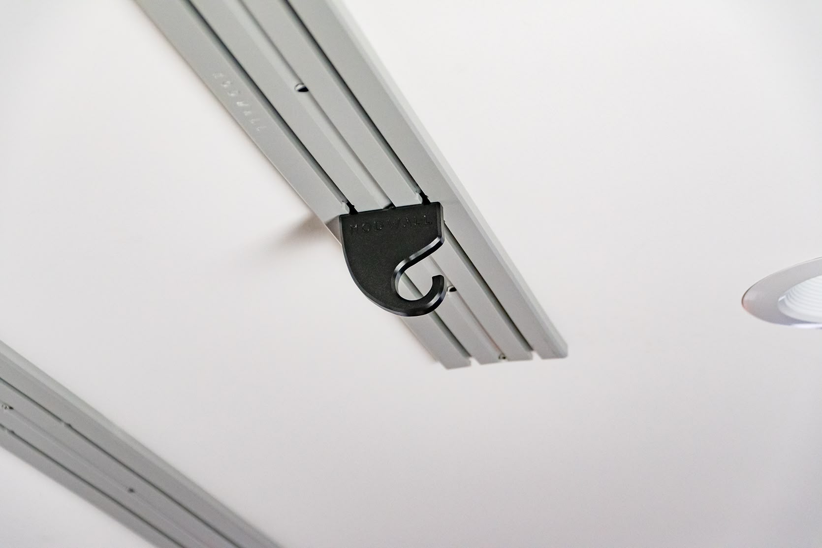 ModWall Ceiling Panel Strip (48″x4″) A close-up view of a black modular hook accessory attached to a light grey ModWall Ceiling Panel Strip mounted on a white ceiling, demonstrating how various accessories can be easily integrated for efficient overhead gear organization.