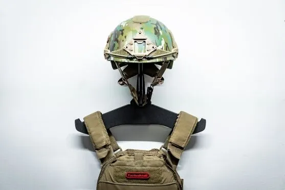 A camo tactical helmet and tactical vest hung on a heavy gear hanger mounted directly to a white wall.