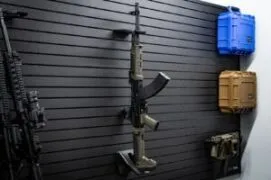 An angled side view of a rifle with a magazine inserted, mounted on a black slatwall system using the ModWall Vertical One Rifle Rack.