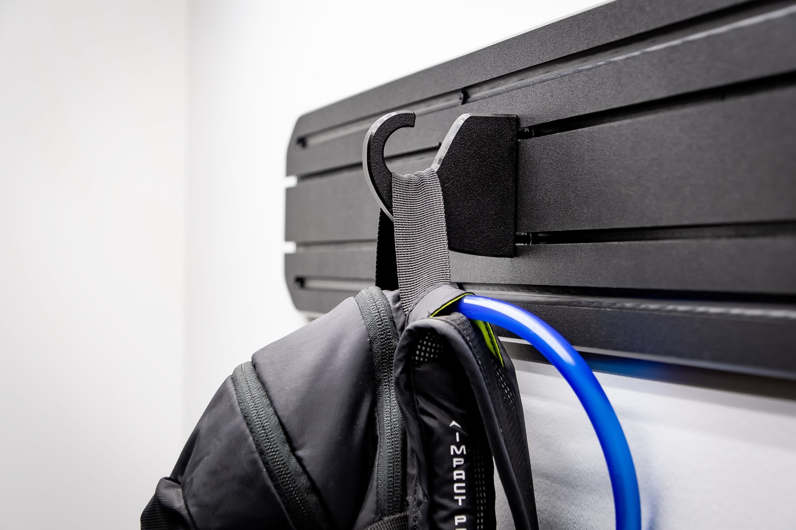 MidMod Panel A close-up, angled view of a black hook attached to a horizontally grooved wall rack, supporting a tactical backpack with a blue hydration tube.