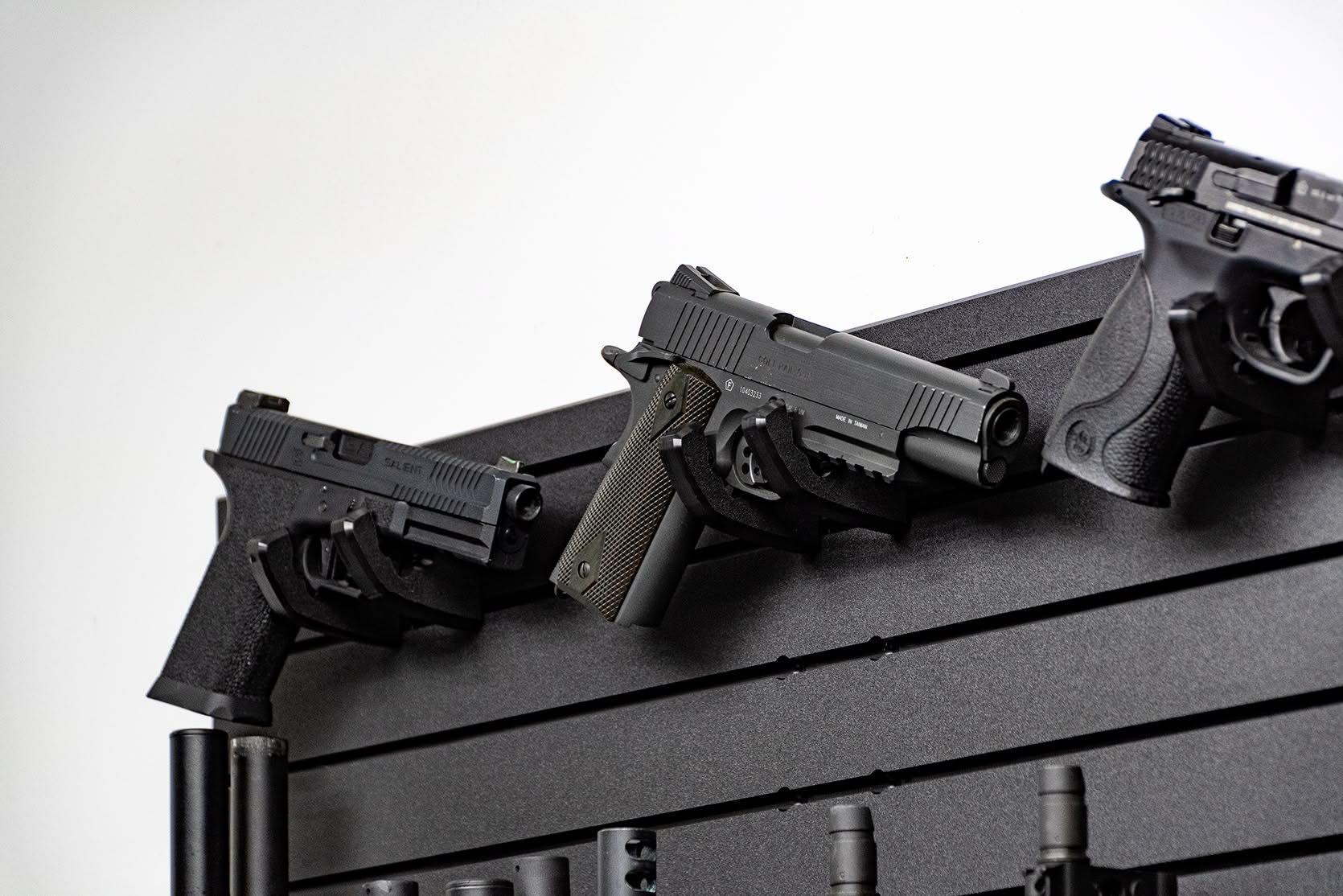 Three handguns mounted at an angle on individual hangers against a black modular wall panel. This display shows how to keep multiple sidearms organized and ready for fast access in a home defense setup.