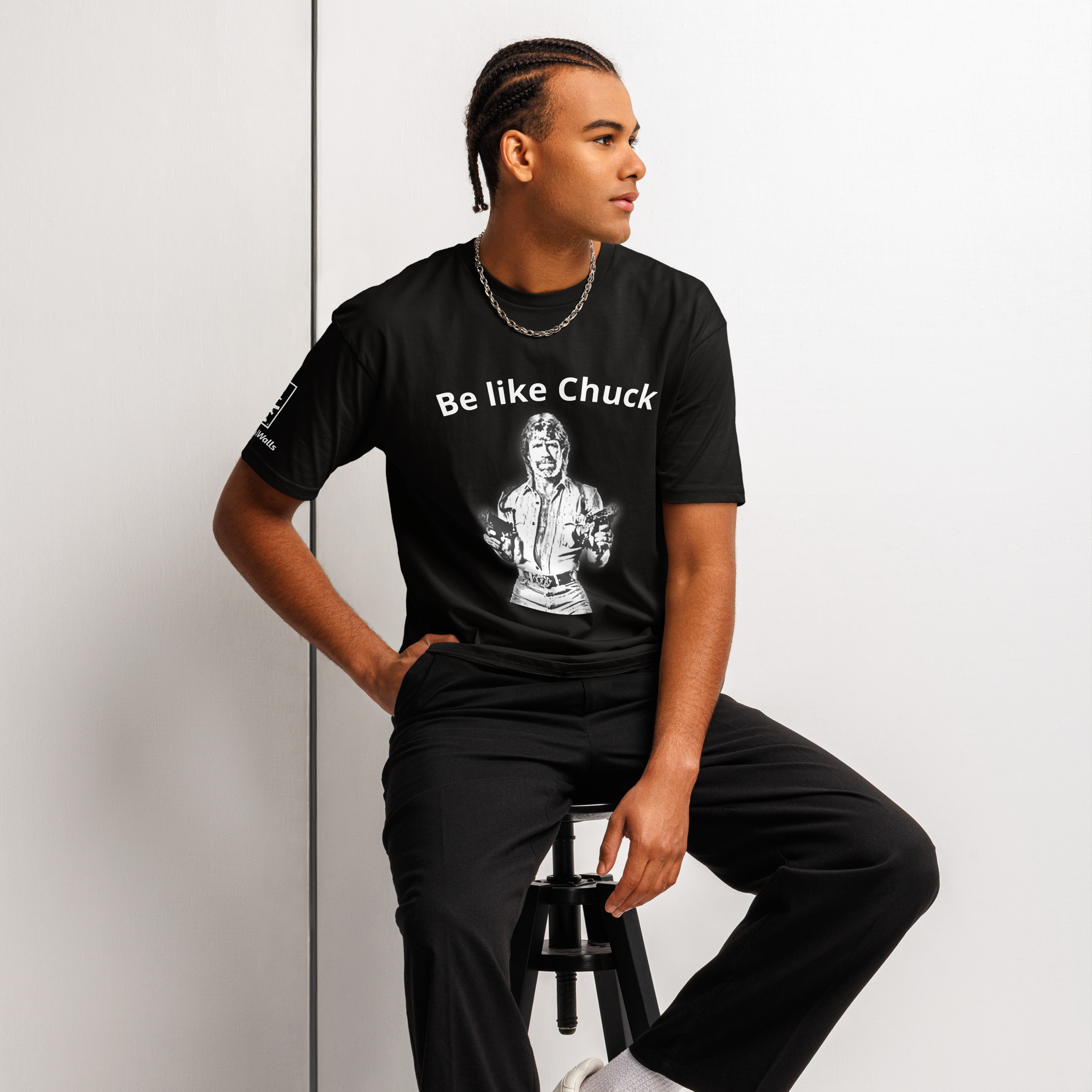 Be Like Chuck T-Shirt Man wearing a black Tactical Walls Be Like Chuck Norris gun shirt with dual-wielding graphic, seated on a stool