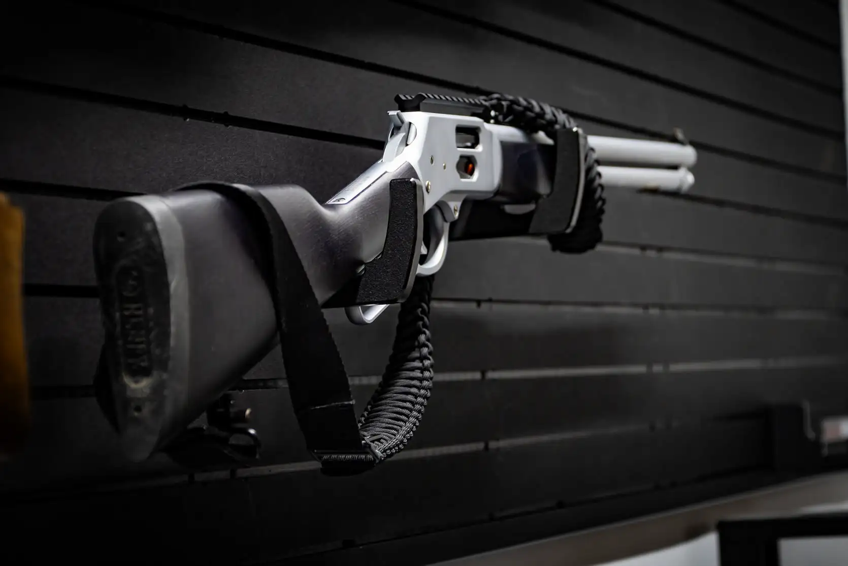 A silver tactical lever-action long gun with a paracord sling resting on sleek black storage wall mounts.
